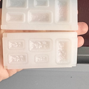 May include: Two clear silicone moulds, each with rectangular and square cavities of varying sizes. The moulds are held in a hand, and the cavities have a textured surface, likely for creating decorative items.