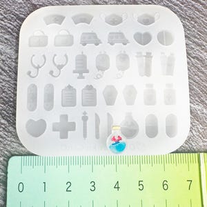 Op de afbeelding: Een doorzichtige siliconen mal met diverse medische vormen, waaronder stethoscopen, kruizen en capsules. Er is ook een kleine, kleurrijke hanger in de vorm van een flesje met een rood en blauw ontwerp. Een liniaal is eronder geplaatst.