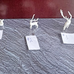 May include: Three small, white reindeer figurines are arranged on a textured gray surface. Each figurine is accompanied by a small white paper with a number written on it. The reindeer are in different poses.