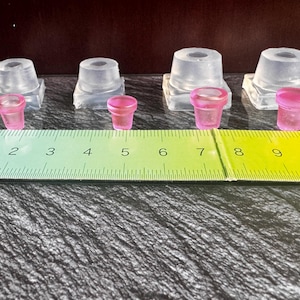 Puede incluir: Cuatro moldes de silicona transparentes y cuatro pequeñas piezas de resina rosa translúcida con forma de maceta dispuestas en fila. Una regla verde y amarilla está colocada frente a los artículos, mostrando la escala de 1 a 10 centímetros.