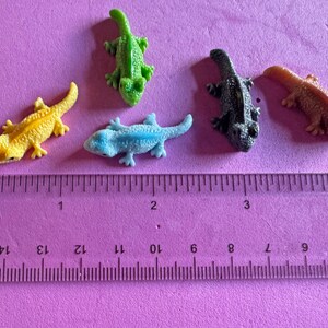 May include: A collection of small, colorful lizard figurines in yellow, green, blue, black, and orange. The lizards are arranged on a pink surface next to a ruler marked in inches.