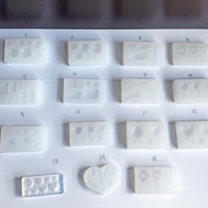 May include: A collection of clear silicone molds in various shapes, including circles, hearts, and geometric designs. The molds are arranged on a white surface, numbered from 1 to 15, and are likely for resin or craft projects.