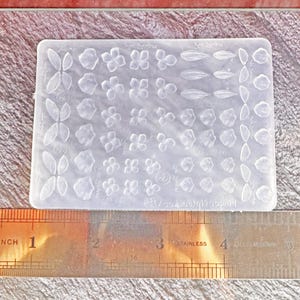 Puede incluir: Un molde de silicona transparente y rectangular con diseños de flores y hojas. Junto al molde, una regla de acero inoxidable muestra medidas en pulgadas. El molde se utiliza para manualidades o fines decorativos.