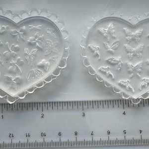 May include: Two clear, heart-shaped molds with butterfly and floral designs. Each mold has a scalloped edge. A ruler with inch measurements is placed below the molds. The molds are approximately 4 inches wide.