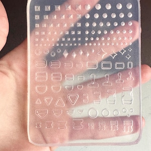 May include: A clear, rectangular silicone mold with various shapes and patterns. The mold features geometric shapes like squares, circles, diamonds, and stars, as well as buckle and strap designs. The mold is held in a hand.