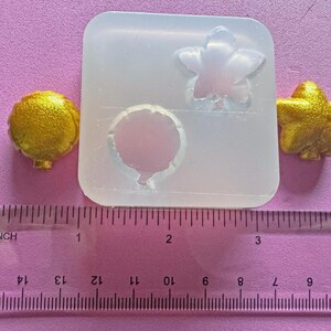 May include: A clear silicone mold with three cavities: a star, a balloon, and a flower. Two gold-colored resin pieces, a star and a balloon, are next to the mold. A ruler with inch measurements is in the foreground.