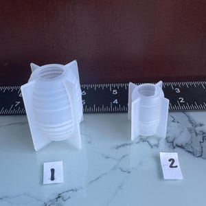 May include: Two translucent white silicone molds shaped like ribbed bombs with fins. The larger mold is labeled with the number 1, and the smaller mold is labeled with the number 2. A black ruler is positioned behind them for scale.