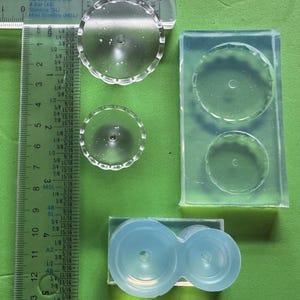 Könnte beinhalten: Klare, kreisförmige Harzformen und fertige Teile werden auf einer grünen Oberfläche gezeigt. Die Formen haben einen gewellten Rand, und ein Lineal ist neben den Objekten platziert. Die fertigen Teile haben einen Durchmesser von etwa 5 cm.