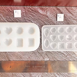 May include: Two clear silicone molds for resin casting, one with various shapes including hearts and squares, and the other with gemstone shapes. A metal ruler with inch measurements is also present.