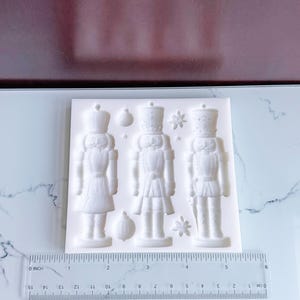 Può includere: Uno stampo in silicone bianco con tre dettagliati disegni di soldati schiaccianoci, con stelle e ornamenti. Lo stampo include un righello per riferimento delle dimensioni, in centimetri.