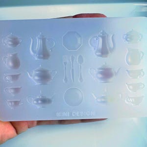 May include: A clear, rectangular silicone mold featuring various miniature tea party elements. The mold includes teapots, teacups, saucers, a plate, and cutlery. The text "MINI DESIGN" is visible at the bottom. Ideal for crafting miniature tea sets.