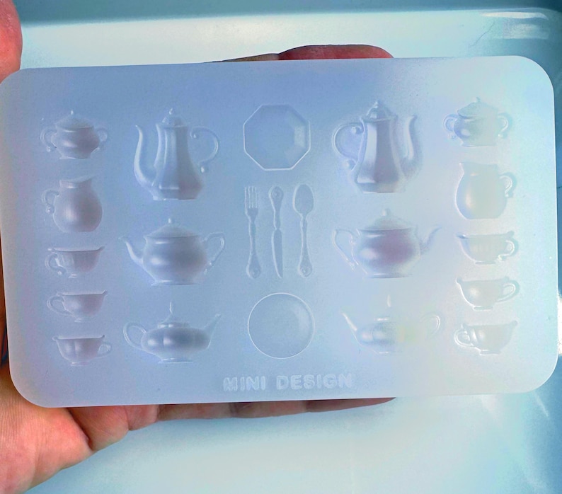 May include: A clear, rectangular silicone mold featuring various miniature tea party elements. The mold includes teapots, teacups, saucers, a plate, and cutlery. The text "MINI DESIGN" is visible at the bottom. Ideal for crafting miniature tea sets.