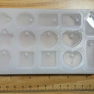 May include: A clear, rectangular silicone mold with various shapes, including a star, heart, and tag designs. The mold is on a wooden surface, with a ruler showing measurements in inches.