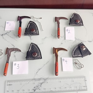 May include: Four miniature hatchets with orange handles and black leather sheaths, labeled #1 through #4. Each hatchet features a unique metal blade finish. A clear plastic ruler at the bottom shows the scale in inches and centimeters.