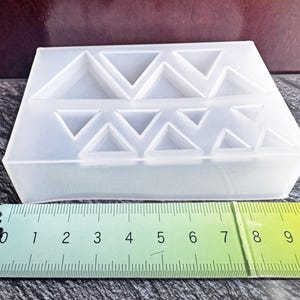 May include: A translucent silicone mold featuring multiple triangular cavities of varying sizes arranged in two rows. A green ruler is positioned in front of the mold, showing a width of approximately 10 centimeters for the entire silicone block.