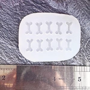 Pode incluir: Um molde de silicone branco com 12 cavidades em forma de osso. Uma régua de metal com medidas em centímetros é colocada abaixo. A régua tem o texto "MADE IN CHINA".