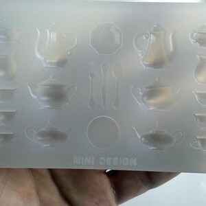 May include: A clear silicone mold featuring miniature designs of teapots, teacups, saucers, and cutlery. The mold includes the text "MINI DESIGN" at the bottom. The designs are arranged in rows and columns.