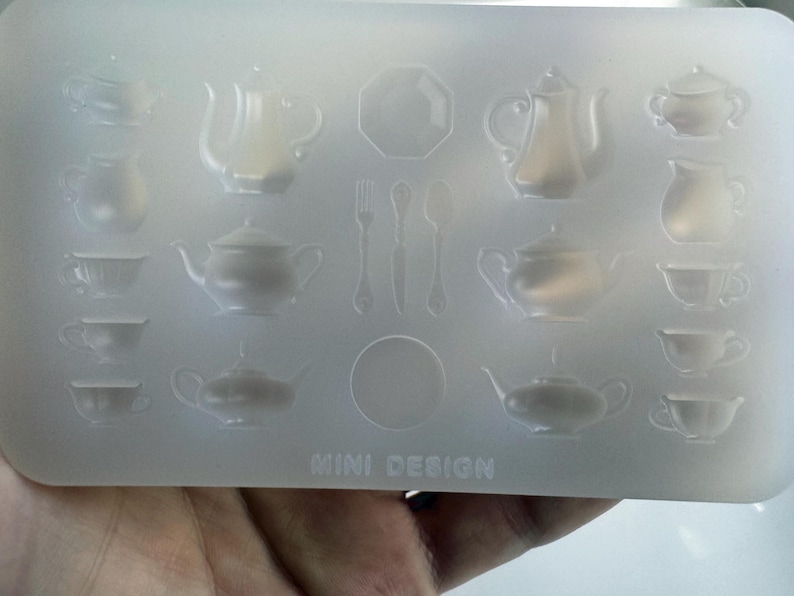 May include: A clear silicone mold featuring miniature designs of teapots, teacups, saucers, and cutlery. The mold includes the text "MINI DESIGN" at the bottom. The designs are arranged in rows and columns.