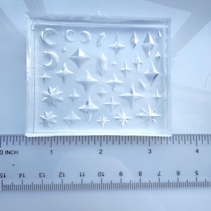 May include: A clear silicone mould featuring a variety of star and crescent moon shapes. The mould includes different sizes and styles of stars, along with several crescent moon designs. The mould is approximately 7.6 cm long and is used for crafting.