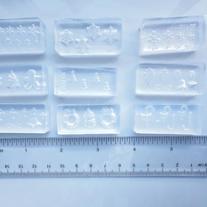 Puede incluir: Nueve moldes rectangulares transparentes con diseños navideños, como copos de nieve, árboles de Navidad y coronas. Una regla con medidas en centímetros en la parte inferior muestra que los moldes miden aproximadamente 2,5 cm de ancho.