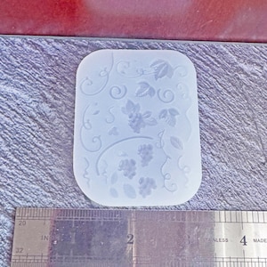 May include: A clear, rectangular silicone mold featuring a grape and vine design. The mold has a detailed pattern of grapes, leaves, and swirling vines. A stainless steel ruler with inch measurements is below the mold.