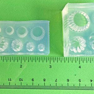 May include: Two clear silicone molds with various circular and floral designs. A ruler with inch measurements is placed on a green surface next to the molds. The molds are for crafting or jewelry making.