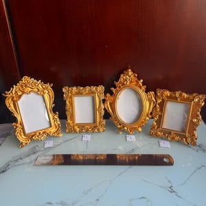 Op de afbeelding: Vier sierlijke, goudkleurige fotolijsten met witte interieurs worden getoond. De frames variëren in vorm, waaronder rechthoekige en ovale ontwerpen. Een liniaal is voor de frames geplaatst. De frames staan op een lichtblauwe en witte ondergrond.