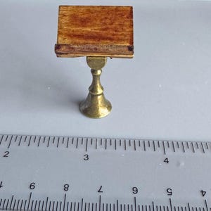 Può includere: Tavolo in miniatura in legno con base a piedistallo color ottone. Il piano del tavolo è rettangolare con finitura a venature naturali. Il tavolo è in scala ridotta, probabilmente per una casa delle bambole o un'esposizione in miniatura.