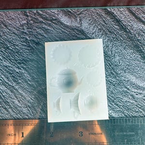 May include: A white silicone mold featuring a teapot, teacups, and decorative circular shapes. The mold is placed on a textured surface with a stainless steel ruler showing measurements in inches.