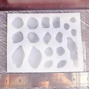 May include: A white silicone mold with various shapes resembling stones. The mold includes a ruler with inch measurements. The shapes vary in size and are arranged in a rectangular pattern. The mold is on a textured surface.
