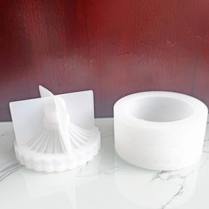 Può includere: Due stampi in silicone bianchi per il crafting. Uno stampo è di forma cilindrica con un motivo decorativo sul lato. L'altro stampo è un design dettagliato a forma di ventaglio con un palo centrale e una base scanalata.