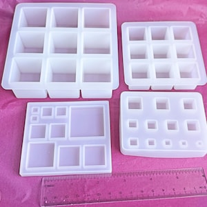 May include: Four clear silicone molds in various square and rectangular shapes, ideal for resin casting. A clear ruler with inch measurements is at the bottom. The molds are arranged on a pink surface.