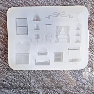 May include: A clear silicone mould with miniature furniture designs. The mould includes shapes of a two-tiered shelf, a lamp, a bow, a window with curtains, a heart-shaped object, a cabinet, a bed, and a window.