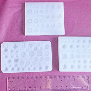 May include: Three white silicone molds with various shapes, including buttons, hearts, stars, and squares, are arranged on a pink surface. A clear ruler with inch measurements is at the bottom. The molds are for crafting.