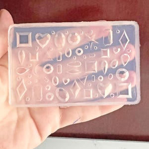 May include: A clear silicone mold filled with various shapes, including squares, hearts, ovals, and diamonds. The mold is rectangular and designed for crafting jewelry or decorative elements. The shapes are arranged in a grid-like pattern.