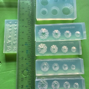 May include: Multiple clear silicone moulds featuring diverse designs such as flowers, shells, and circular patterns. A green ruler with centimetre and inch markings is positioned beside the moulds. These moulds are for crafting.
