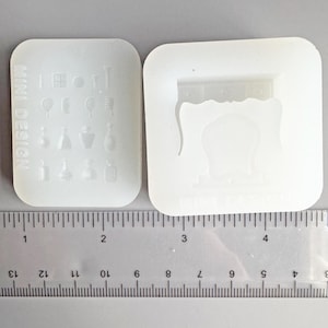 Puede incluir: Dos moldes de silicona blancos para manualidades. Un molde presenta miniaturas de frascos de perfume y otros artículos cosméticos. El segundo molde tiene un diseño de muebles decorativos. Una regla se coloca debajo, con medidas en centímetros.