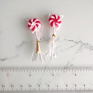 May include: Two red and white swirled lollipops in clear cellophane wrappers. The lollipops are on white sticks, with gold twist ties. A ruler is in the foreground.