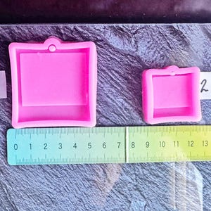 May include: Two pink square silicone molds with a small hole at the top for hanging. The larger mold measures approximately 2.75 inches wide, and the smaller mold measures approximately 1.5 inches wide, as shown by the green ruler placed below them.