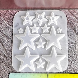 Puede incluir: Un molde de silicona translúcido con varias cavidades en forma de estrella de diferentes tamaños. Una regla amarilla está colocada en la parte inferior, mostrando que el molde mide aproximadamente 13 centímetros de ancho. Contiene once huecos.