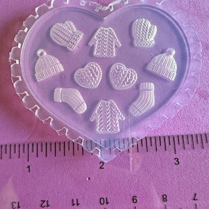 May include: A clear, heart-shaped mould featuring miniature winter-themed designs. The mould includes tiny mittens, sweaters, hats, socks, and heart shapes. A ruler is placed below the mould, with measurements in centimetres.