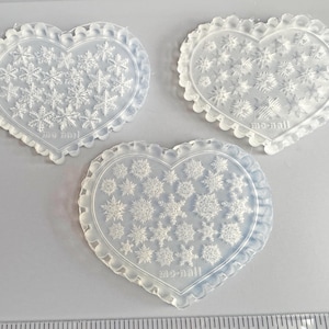 May include: Three clear, heart-shaped molds with snowflake designs. Each mold has a scalloped edge and the text "mo-nail" at the bottom. The molds are made of a translucent material and are arranged on a light surface.