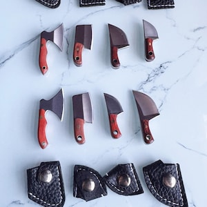 May include: A selection of miniature axes and knives. The axes have red handles, the knives have brown handles, and all have metal blades. Each blade is paired with a black leather sheath with a silver snap closure. The blades are displayed on a marble surface.