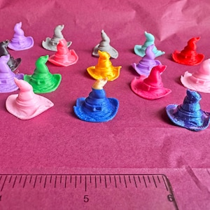 Puede incluir: Una colección de pequeños sombreros de bruja en varios colores, como rosa, verde, azul, amarillo y rojo. Los sombreros están colocados sobre una superficie rosa, con una regla en la parte inferior. Estos sombreros en miniatura son ideales para manualidades o decoración.
