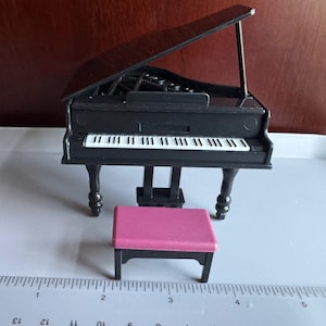 Puede incluir: Un piano de cola negro en miniatura con un banco rosa. El piano tiene teclas blancas y negras y se apoya sobre cuatro patas curvas. El banco es rectangular y tiene cuatro patas negras. El piano mide unos 10 cm de largo.