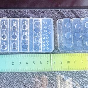 Può includere: Due stampi in silicone trasparente per colata di resina. Uno stampo presenta varie forme di bottiglie, cuori, stelle e altri disegni. Il secondo stampo ha cerchi, cuori e quadrati. Un righello verde è posto sotto, con misure in centimetri.