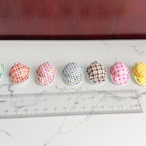 Peut inclure: Sept casquettes de style gavroche miniatures à carreaux de différentes couleurs, dont turquoise, rouge, rose, bleu, marron et jaune. Une règle transparente avec des marquages en centimètres est placée en dessous, servant de référence de taille. Les casquettes sont disposées sur une surface blanche.