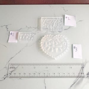 May include: Clear resin molds in various shapes, including rectangular and heart-shaped designs, with seashell and ocean-themed patterns. A clear ruler with centimetre measurements is also present. The molds are labeled with numbers on small white tags.