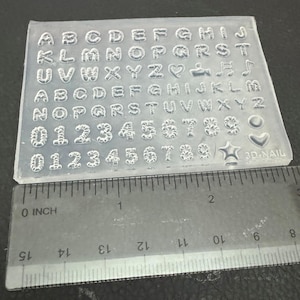 May include: Clear silicone mold with letters, numbers, and symbols for nail art. Includes the alphabet, numbers 0-9, musical notes, hearts, and a star. A ruler is placed next to the mold for scale.
