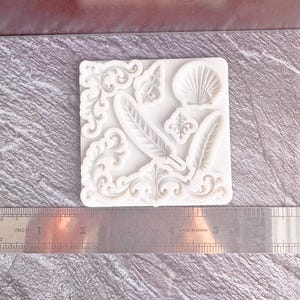Puede incluir: Un molde de silicona blanco con elementos decorativos. El molde incluye dos plumas, una concha y adornos ornamentados. Una regla con medidas en pulgadas es visible debajo.